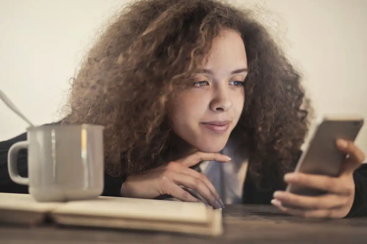 Tânără zâmbitoare care derulează pe telefon aplicațiile, cu o cană de cafea alături