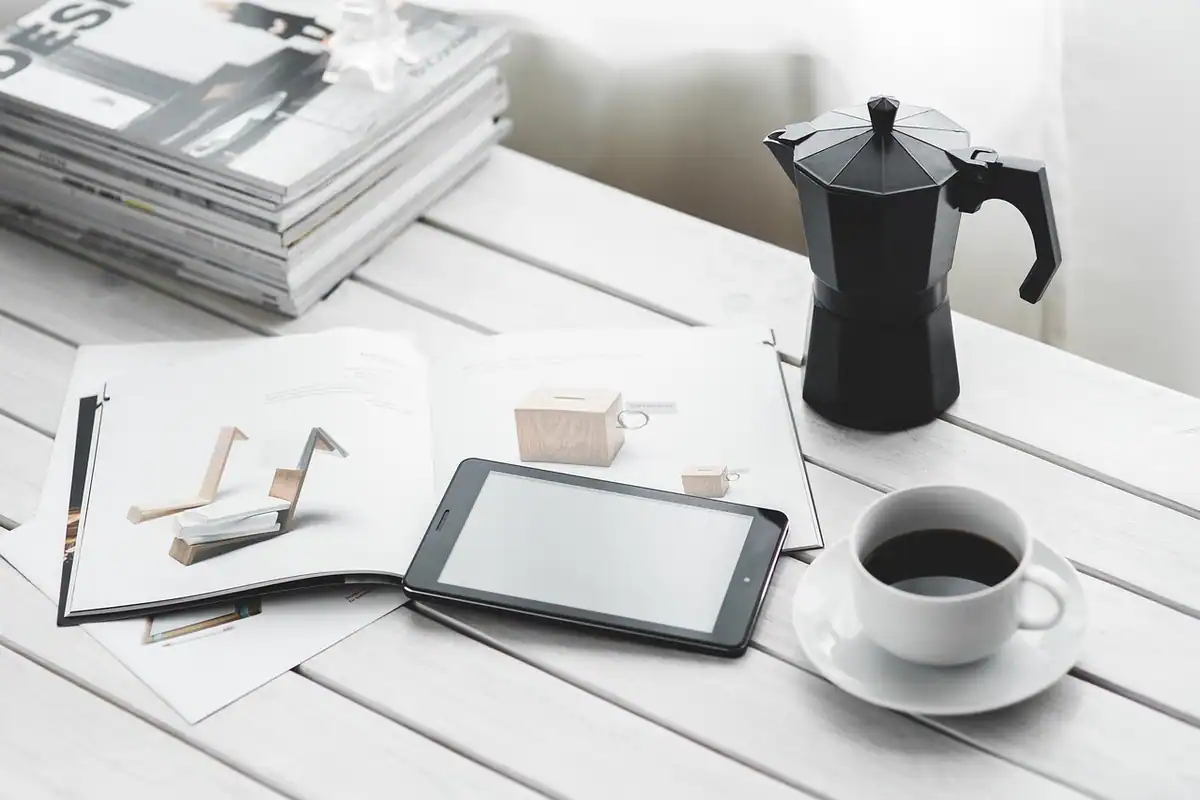 Masă albă cu reviste, tabletă, ibric negru și o ceașcă de cafea