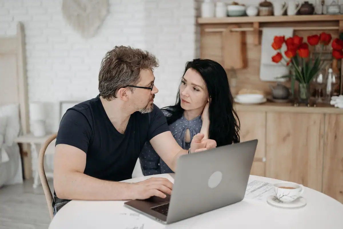 Cuplu care discută în bucătărie, în fața unui laptop deschis pe masă