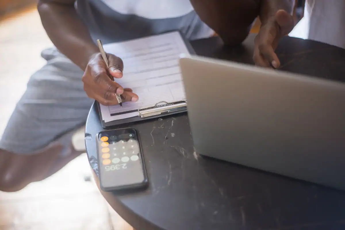 Persoană care completează o foaie pe clipboard, lângă un laptop și un calculator