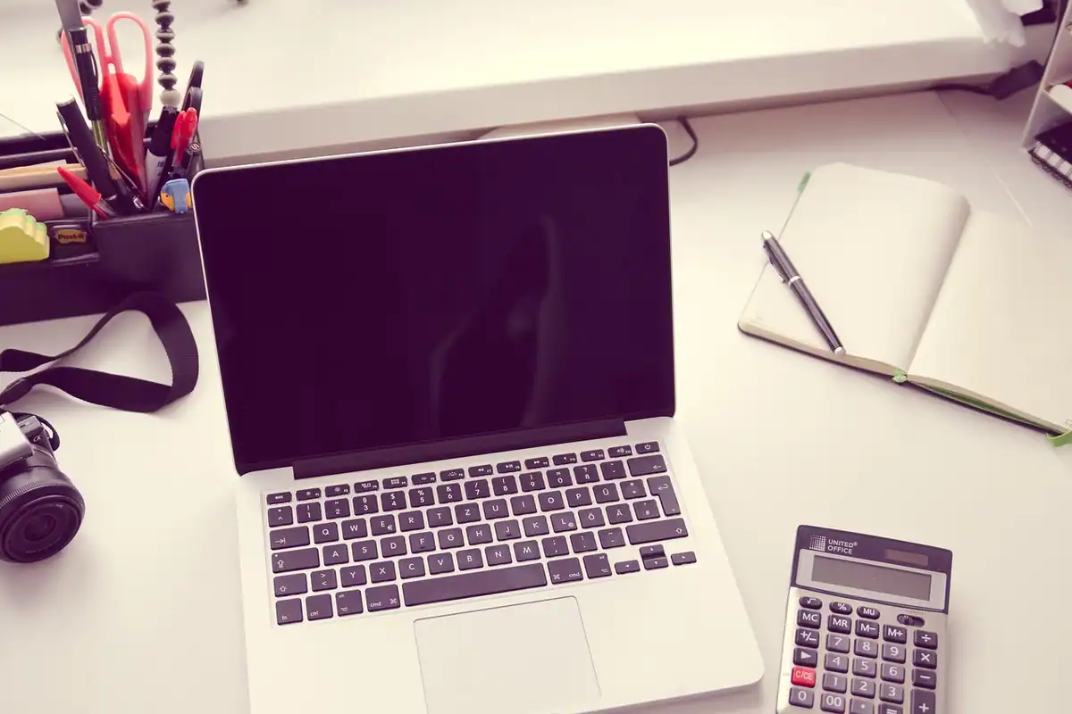 Laptop deschis, caiet cu pix și un calculator mic pe un birou alb