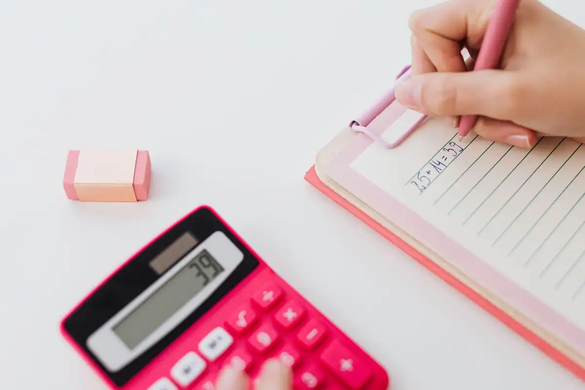 Mână care scrie pe un clipboard roz, lângă un calculator roșu